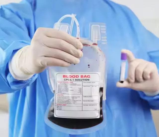 A technician in protective clothing holding a unit of whole blood and a crossmatch sample